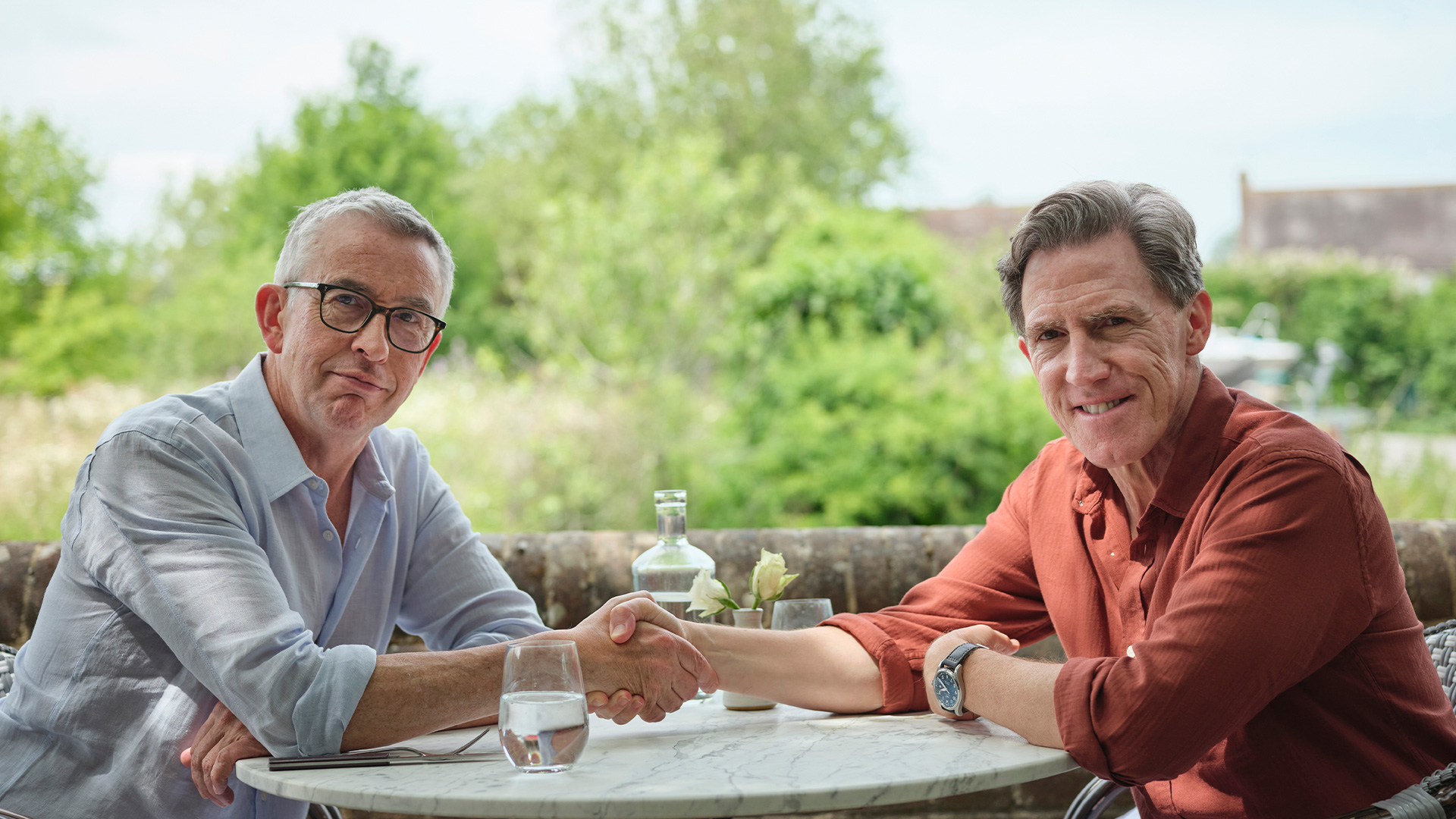 Steve Coogan and Rob Brydon shake hands across an outdoor table in The Trip publicity photograph.